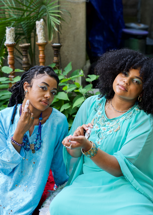 Two female models in a matching blue aesthetic are applying the female on to their skin. One model applies the perfume onto her neck while other applies it on her wrist